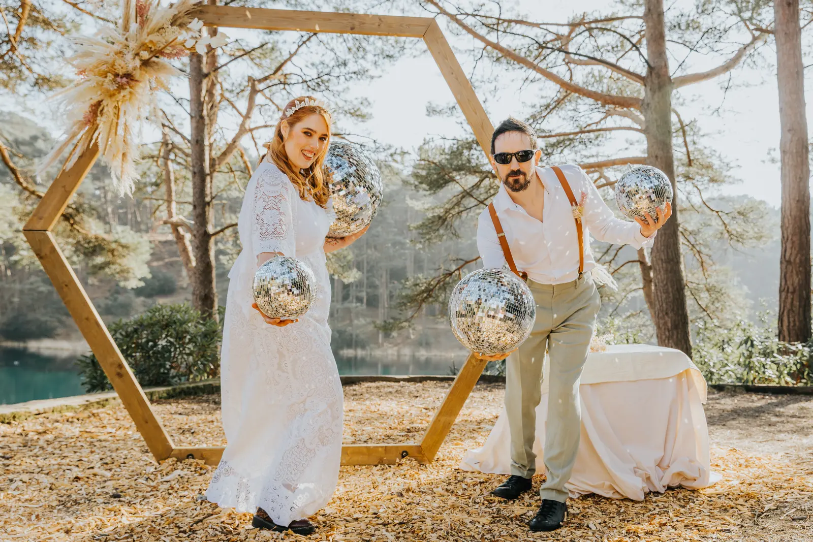 A couple dance with disco balls in their hands in front of the arch, grinning, the lake bright behind them.